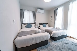 Une chambre d’hôtel moderne avec deux lits, une grande fenêtre avec des rideaux, un appareil de climatisation et une petite table de chevet avec une lampe.