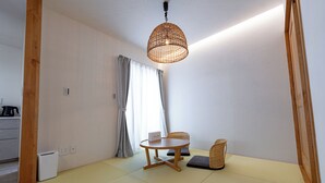A room with tatami flooring, a round table with a cushion, a woven hanging light fixture, and a view of a garden through a window with curtains. - GUEL PALACIO (Miyakojima)