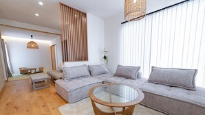 A modern living room with a sectional sofa, a glass coffee table, wooden flooring, and a large window with blinds. - GUEL PALACIO (Miyakojima)