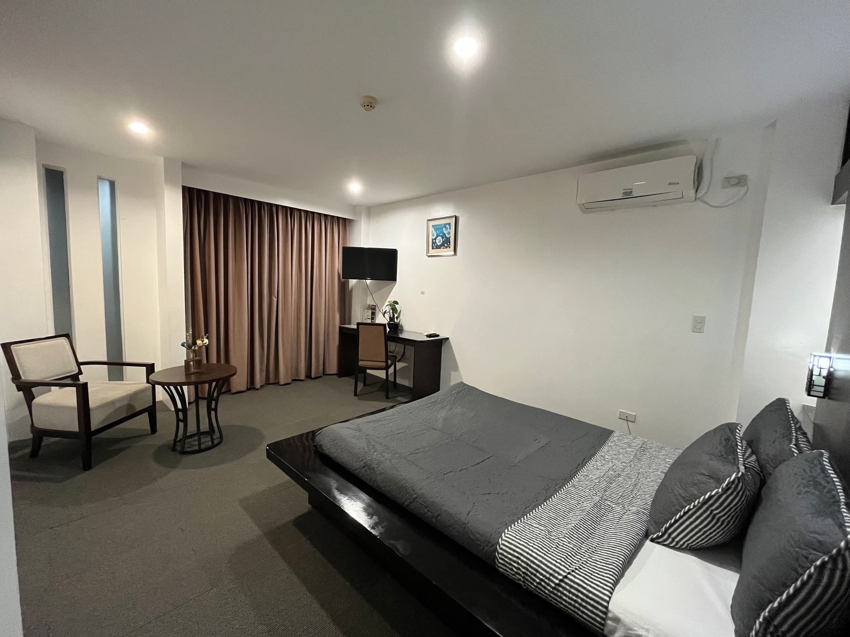 Habitación de hotel con cama, escritorio, silla, televisor y ventana con cortinas.
