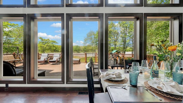 A dining area with a view of an outdoor deck, large windows, and a table set with blue glassware and a bottle. - Lakefront Home in the Poconos with Hot Tub! (Albrightsville)