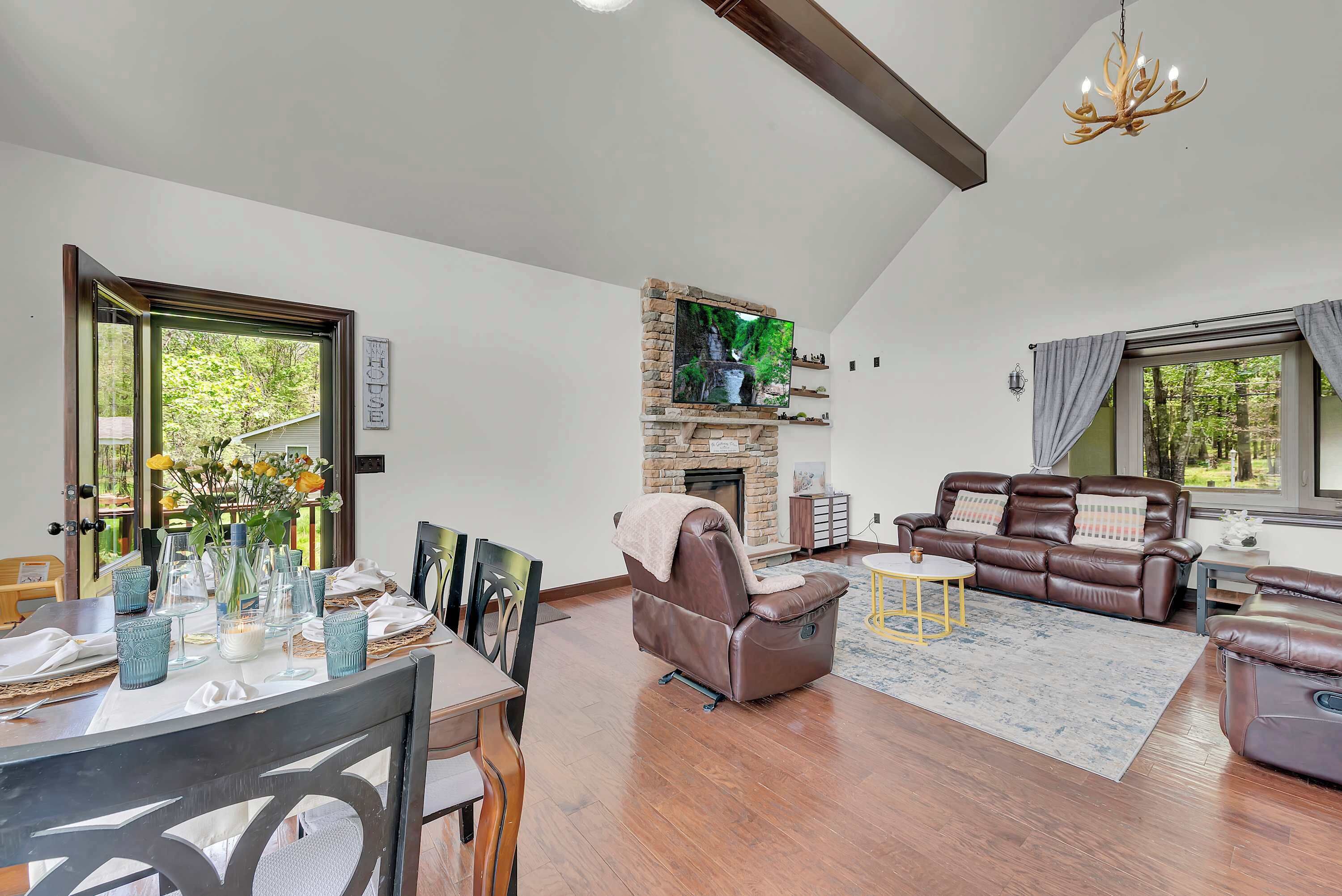 A spacious living room with a fireplace, leather sofas, a dining area with a table set for two, and a large window with a view of trees.