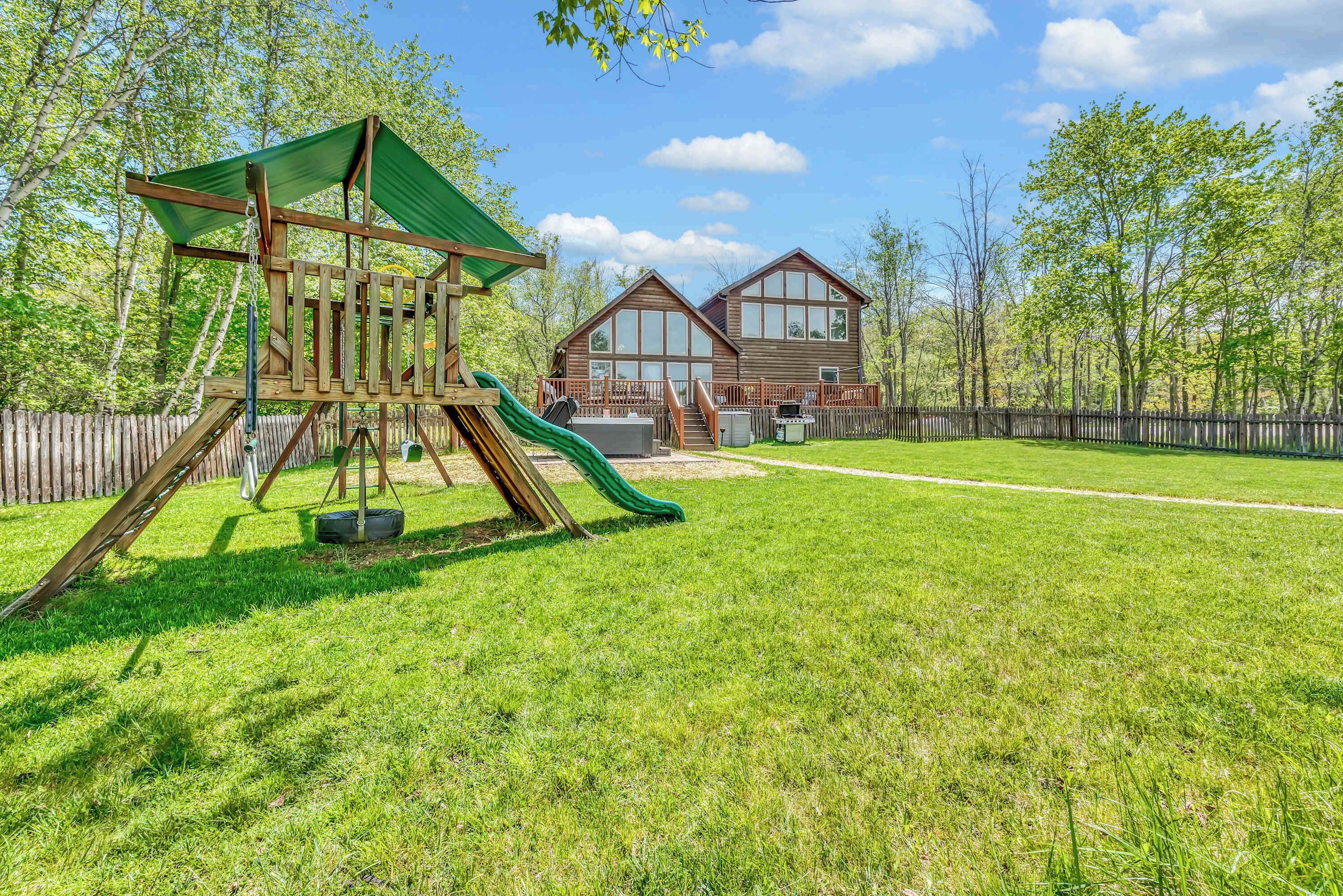 A wooden play structure with a slide and a swing, a grassy yard, and a two-story house with large windows.