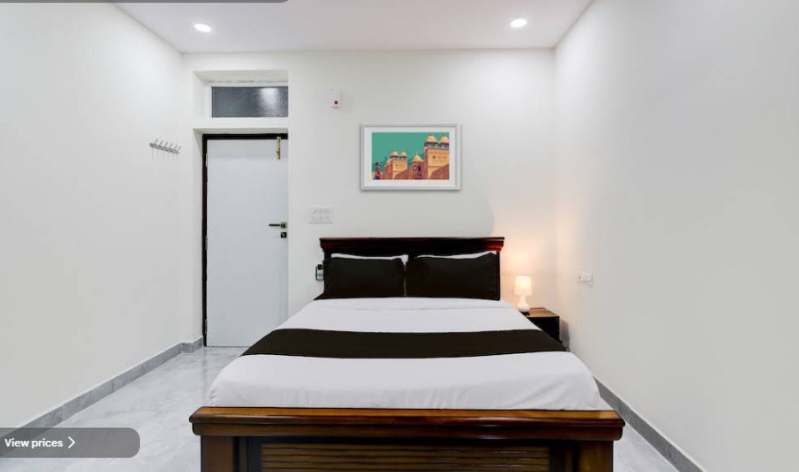A hotel room with a bed, a nightstand, a wall-mounted painting, a door, and a small window.
