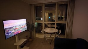 A living room with a TV on the wall, a dining table, and a view of a cityscape through large windows.