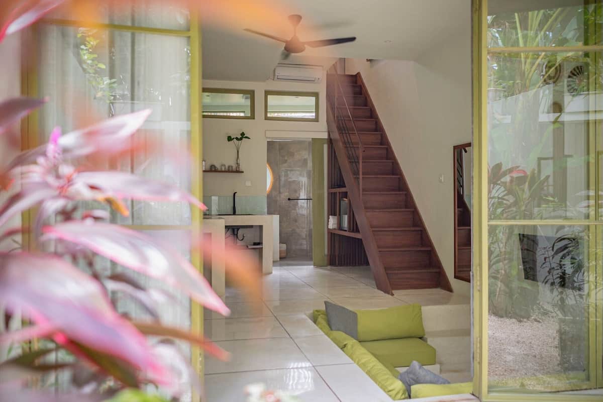 Una sala de estar moderna con una escalera, un comedor y vistas a un jardín a través de amplios ventanales.