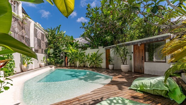 Zona de piscina con terraza de madera, rodeada de exuberante vegetación y un edificio blanco con tejado de baldosas.