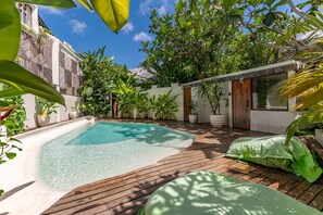Área de piscina com deck de madeira, cercada por vegetação exuberante e um prédio branco com telhado telhado.