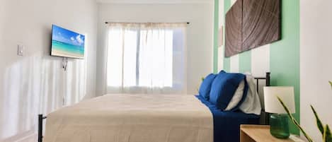 Une chambre à coucher avec un lit, un téléviseur fixé au mur, une fenêtre avec des rideaux et une table de chevet avec une lampe.