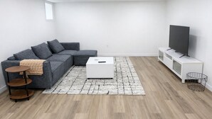 Un salon moderne avec un canapé d’angle gris, une table basse blanche, une petite table d’appoint en bois, un téléviseur posé sur un meuble blanc et un tapis à motifs.