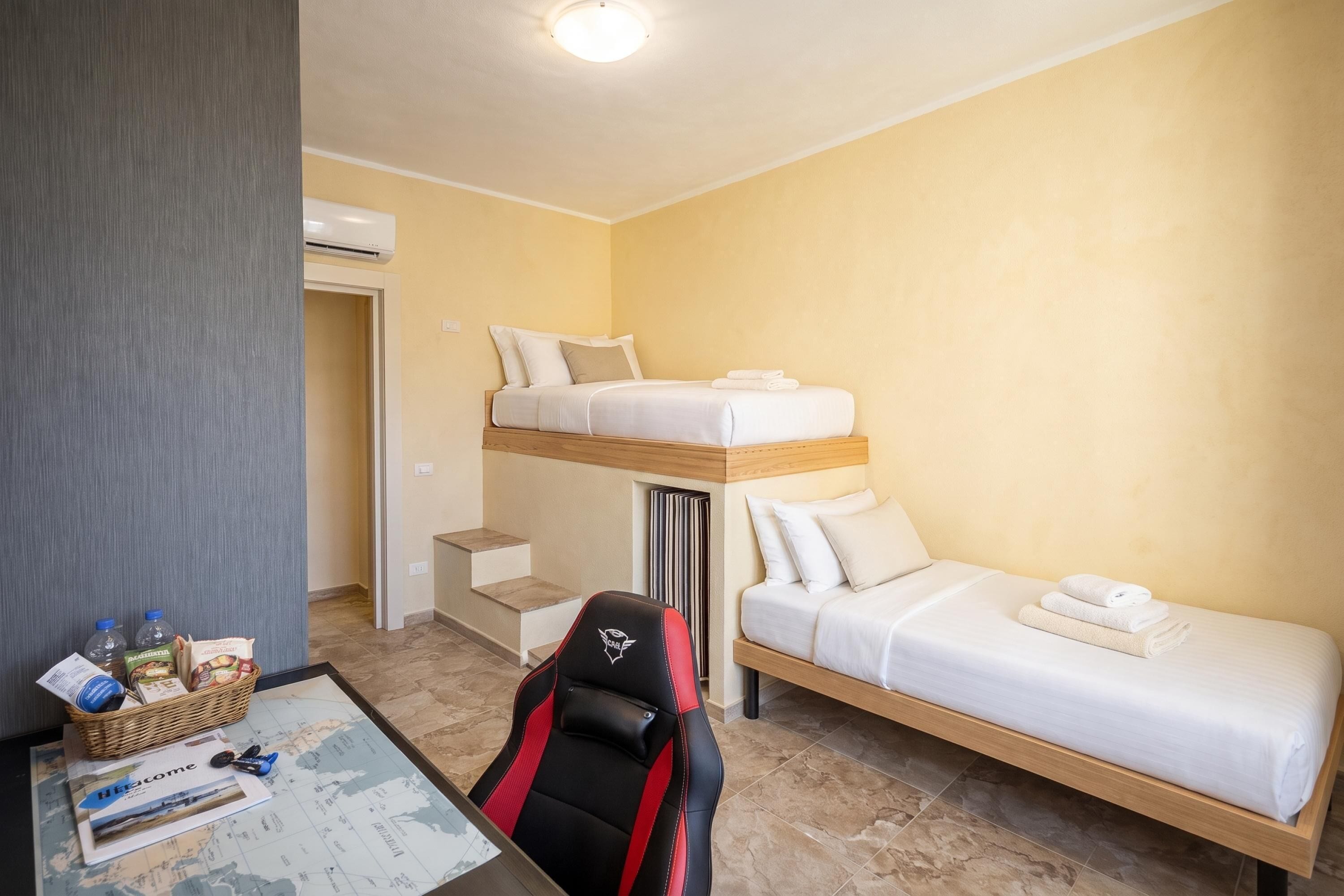 Ein Hotelzimmer mit einem Etagenbett, einem Einzelbett, einem Schreibtisch mit Stuhl und einem kleinen Tisch mit Gegenständen darauf.