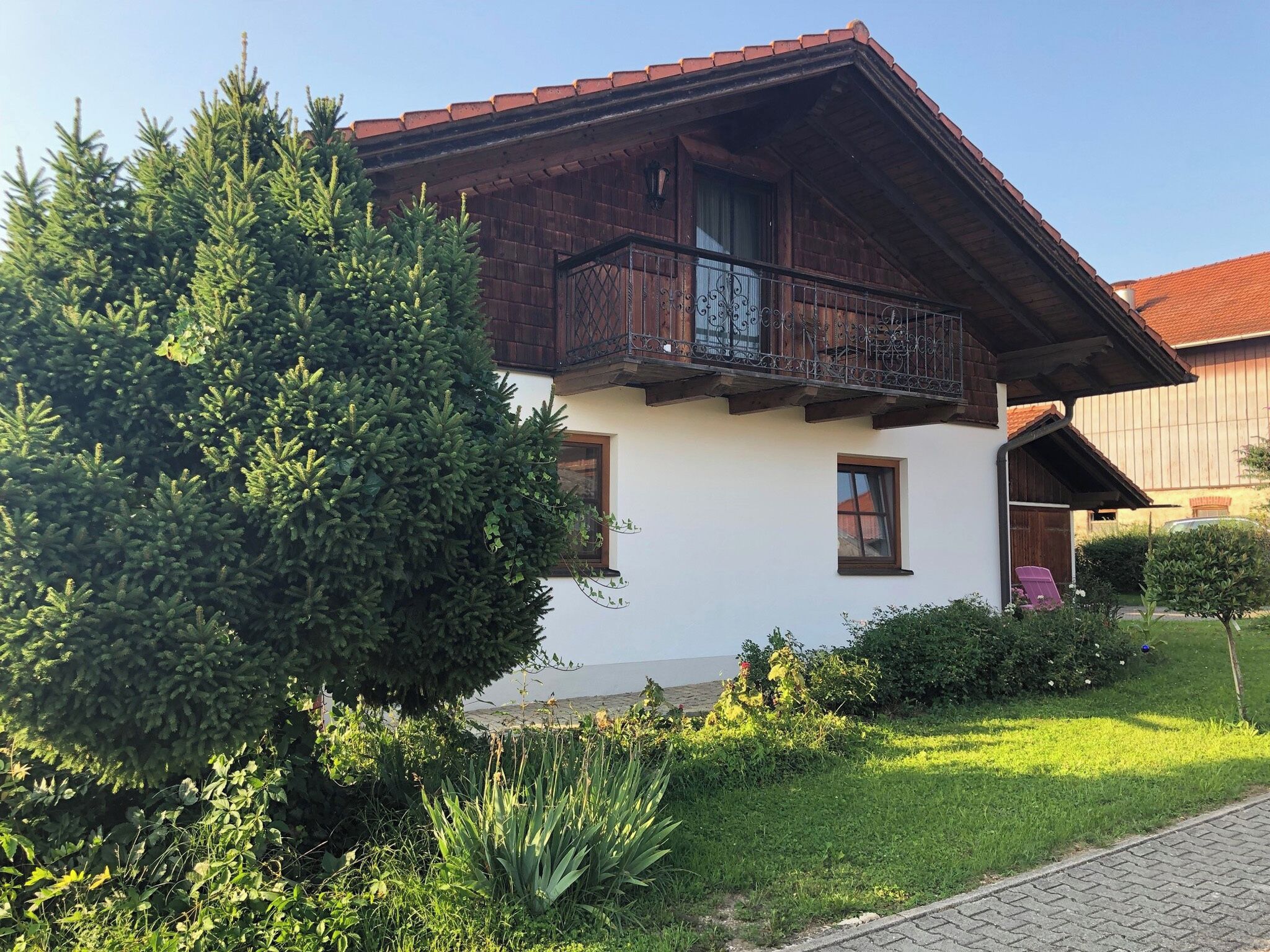 Een tweelaags huis met een houten balkon, omgeven door groen en een verharde oprit.