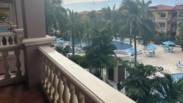 Un balcón de hotel con vista a un área de piscina, palmeras y un edificio con tejados rojos.