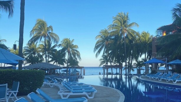 Un espace aménagé au bord de la piscine, avec des chaises longues et des parasols, des palmiers et une vue sur la mer.