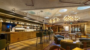 Een moderne bar met een houten toonbank, barkrukken en een zithoek met tafels en stoelen.
