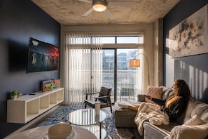 A modern living room with a large TV, a sofa, a coffee table, and a view of the city through large windows. - Chic Downtown Phoenix Gem w/ Scenic Balcony & Park (Phoenix)