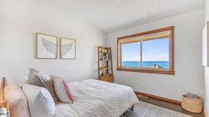 A bedroom with a bed, a window with a view of the sea, and two framed artworks on the wall. - Sea to Sky Retreat by Manzanita Beach Getaway: Ocean Views, 3BR Hilltop Home (Nehalem)