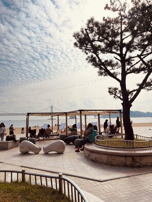 A covered seating area with stone benches and a wooden pergola, overlooking a beach and a bridge.