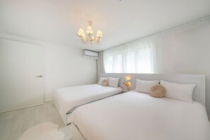 A hotel room with two beds, a chandelier, an air conditioning unit, and a window with sheer curtains.