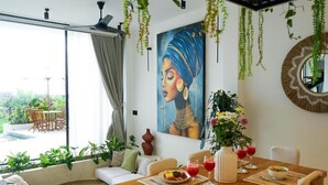 A dining area with a wooden table set for two, a painting on the wall, and hanging plants. - Dzen Greenfield Villa 9 (Ubud)