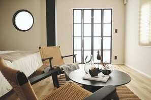 Una habitación acogedora con cama, una mesa redonda con un libro y un florero, una silla de mimbre y un ventanal con persianas.