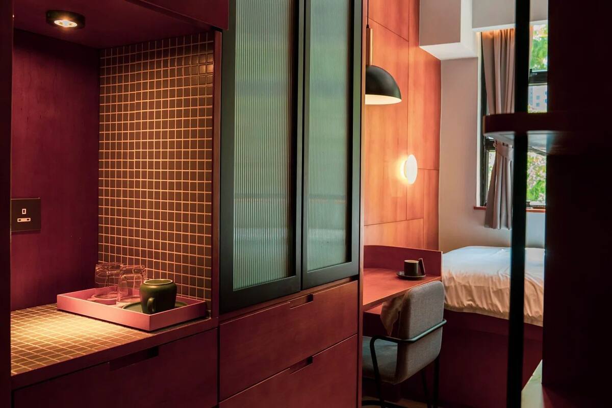 Una habitación de hotel compacta con cama, escritorio con silla, ventana con cortinas y una lámpara de pared.
