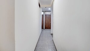 A narrow hallway with white walls and a grey tiled floor, leading to a door with a wooden panel design. - Roomshala Hotel In Jangpura (New Delhi)