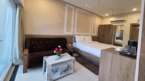 A hotel room with a bed, a sofa, a coffee table with flowers, a desk, a mirror, and an air conditioning unit. - The Manthan Hotel - City View (Faizabad)