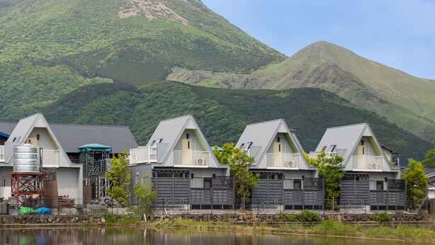 A row of modern, grey-roofed cabins with balconies, set against a backdrop of lush green mountains and a calm lake. - Yufuin Onsen 8A1 Yaeichi (Yufu)