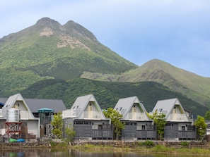 Una hilera de cabañas modernas con techos grises y balcones, con vistas a montañas verdes y un lago tranquilo.