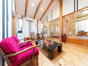 Una habitación con piso de madera, una mesa negra y una silla rosa. Se observan vigas de madera en el techo y un panel de madera con diseño de celosía.