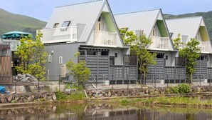 A row of modern, grey buildings with balconies, a calm water body in the foreground, and a hilly landscape in the background. - Yufuin Onsen 8A1 Yaeichi (Yufu)