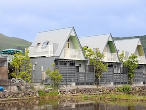 A row of modern, grey buildings with balconies, a calm water body in the foreground, and a hilly landscape in the background. - Yufuin Onsen 8A1 Yaeichi (Yufu)