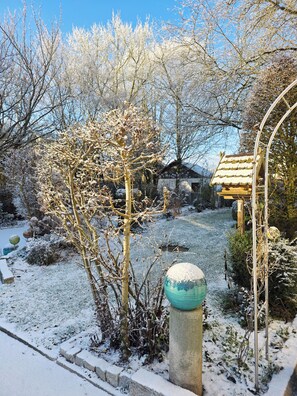 A snow-covered garden with a wooden gazebo, a stone pillar, and a small house in the background.