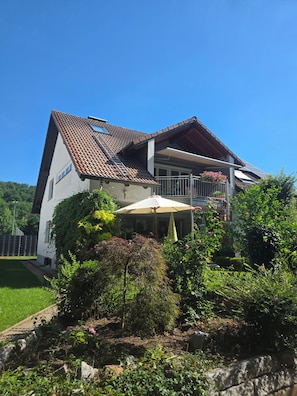 A two-story house with a sloped roof, a balcony with potted plants, and a garden area with various shrubs and flowers.