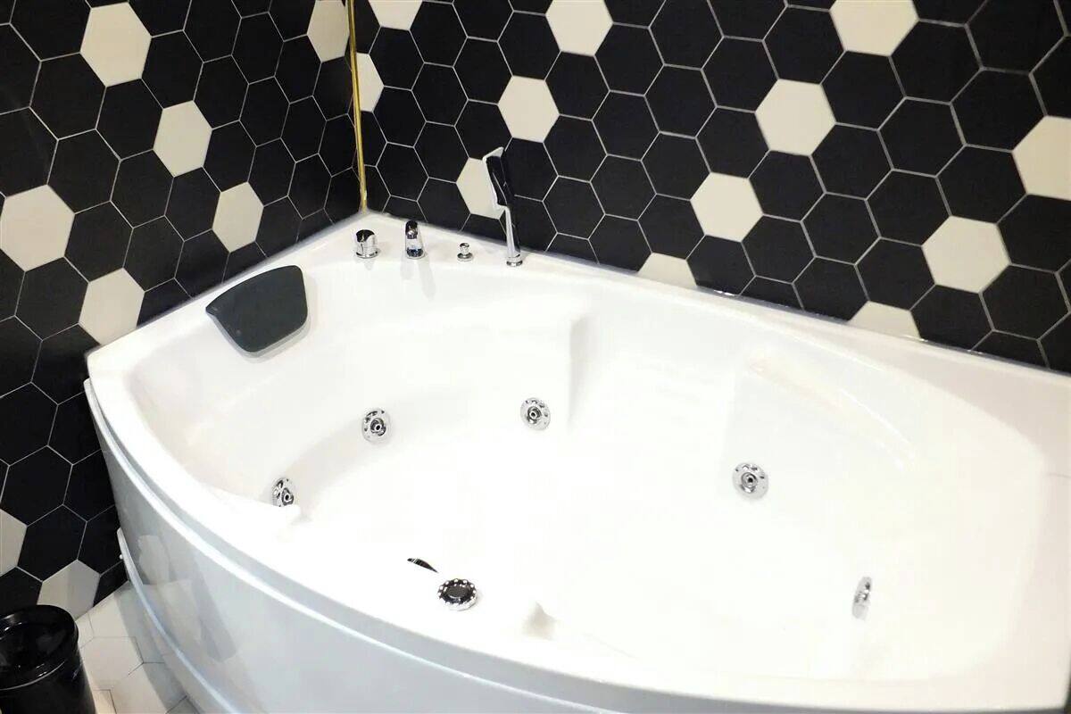 Une baignoire blanche, dotée de plusieurs robinets et d’un drain, est placée devant un mur recouvert de carreaux hexagonaux noirs et blancs.