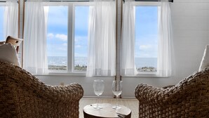 Une pièce avec vue sur la mer, grâce à de grandes fenêtres, deux fauteuils en osier et deux verres à vin vides sur une table en bois.