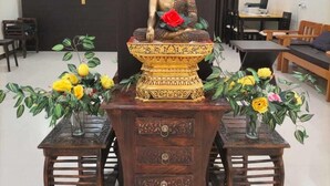 A statue of a seated figure with a red flower, surrounded by two wooden tables with floral arrangements. - Hotel Temple View (Gaya)