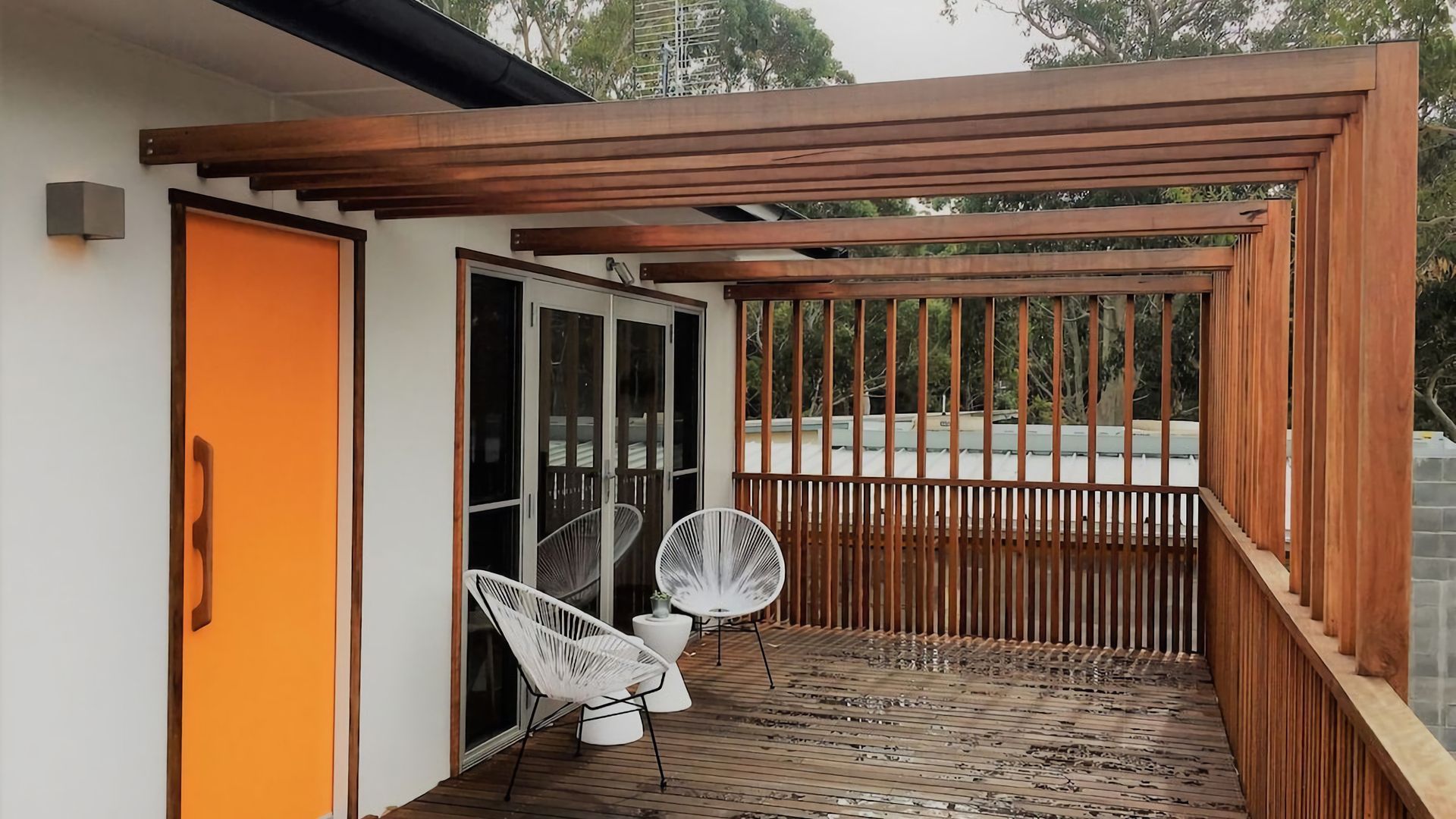 A wooden deck with a pergola, two white chairs, and a sliding glass door.