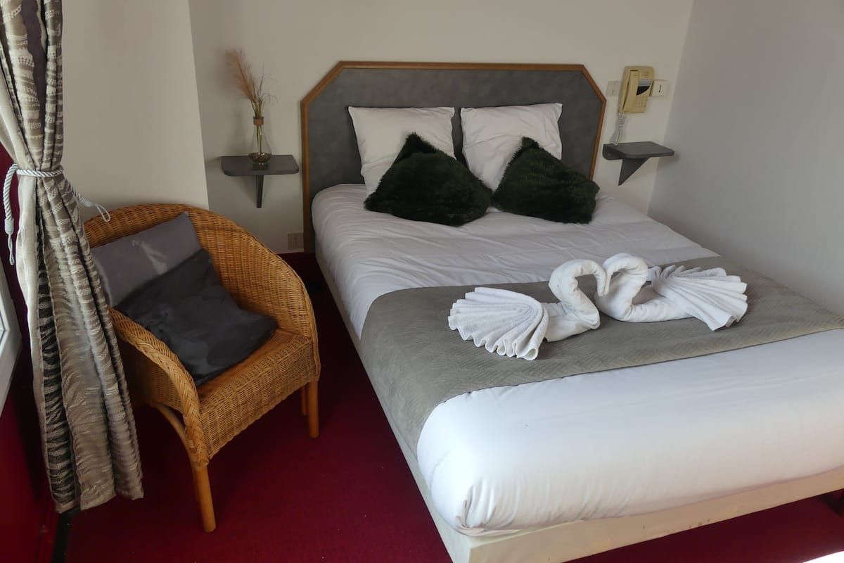 A hotel room with a bed, a wicker chair, a bedside table with a vase, and a wall-mounted phone.