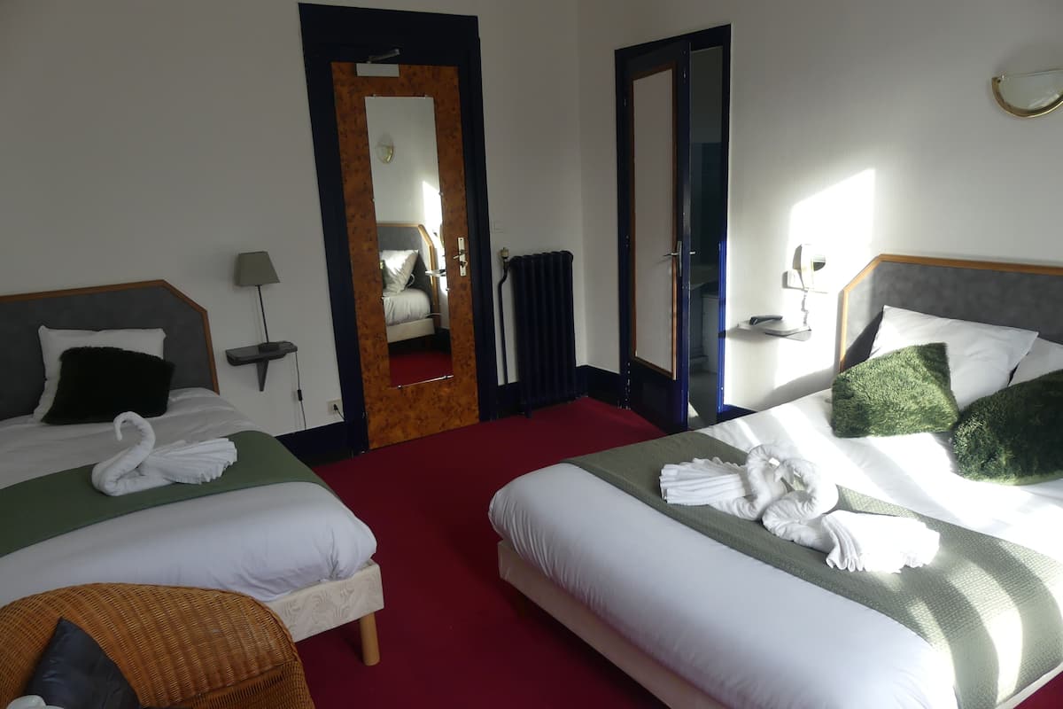 A hotel room with two beds, a red carpet, a wicker chair, a table with a remote control, and a mirror.