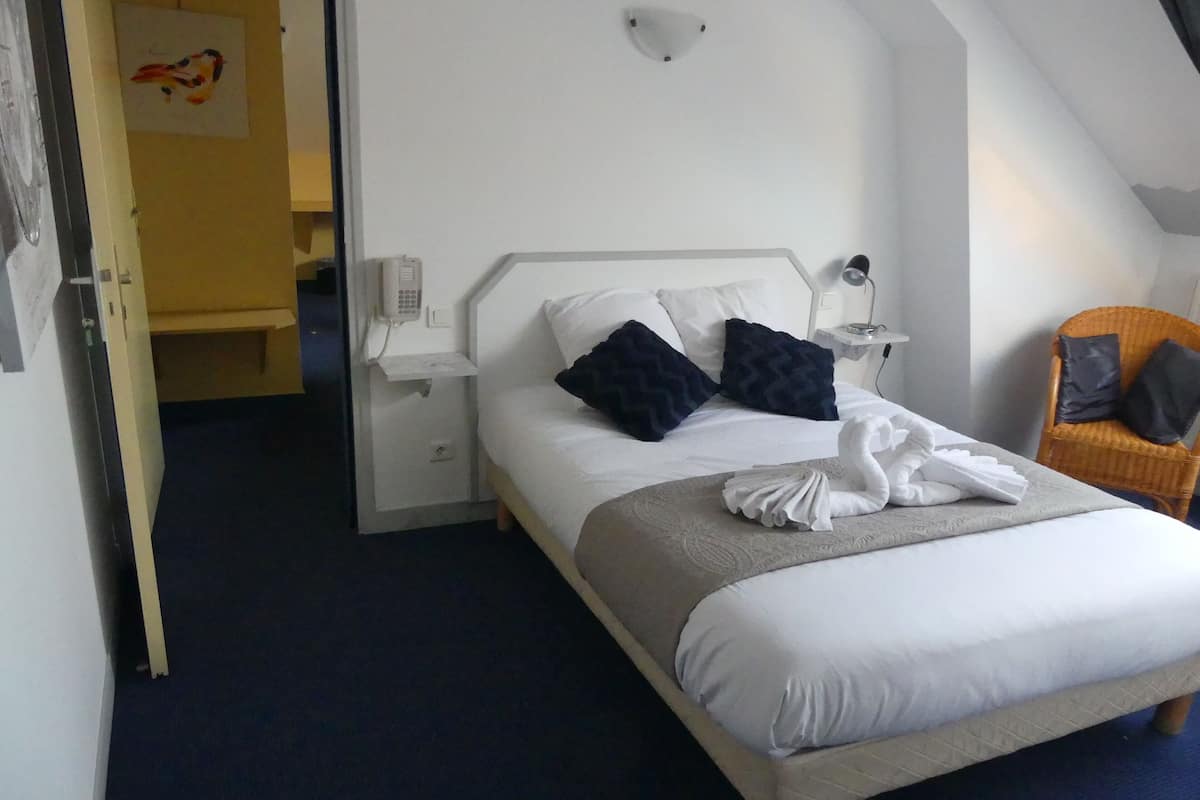 A neatly made bed with white linens and two dark pillows, a bedside table with a lamp, and a chair with a cushion.