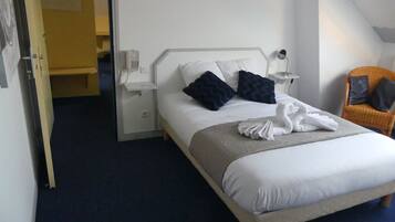 A neatly made bed with white linens and two dark pillows, a bedside table with a lamp, and a chair with a cushion.