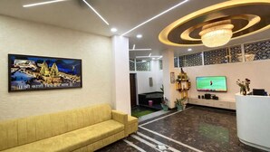 A hotel lobby with a reception desk, a television, a framed picture of a temple, a yellow sofa, and a chandelier. - Room Racers (Varanasi)