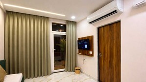 A modern hotel room with a sliding glass door, a flat-screen TV mounted on the wall, a wooden door, and a marble floor. - Room Racers (Varanasi)