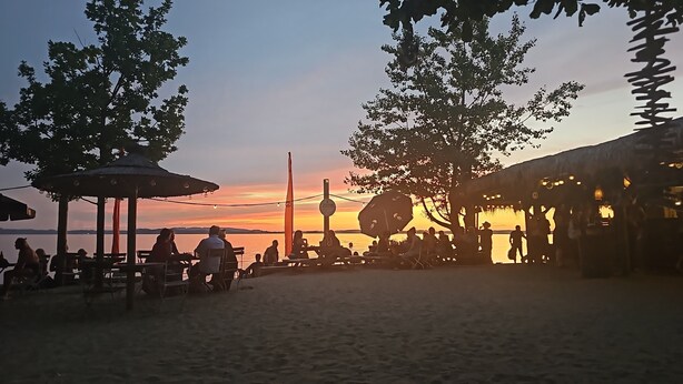 Um cenário à beira-mar com pessoas sentadas em mesas sob guarda-sóis, uma estrutura com telhado de palha e um pôr do sol ao fundo.