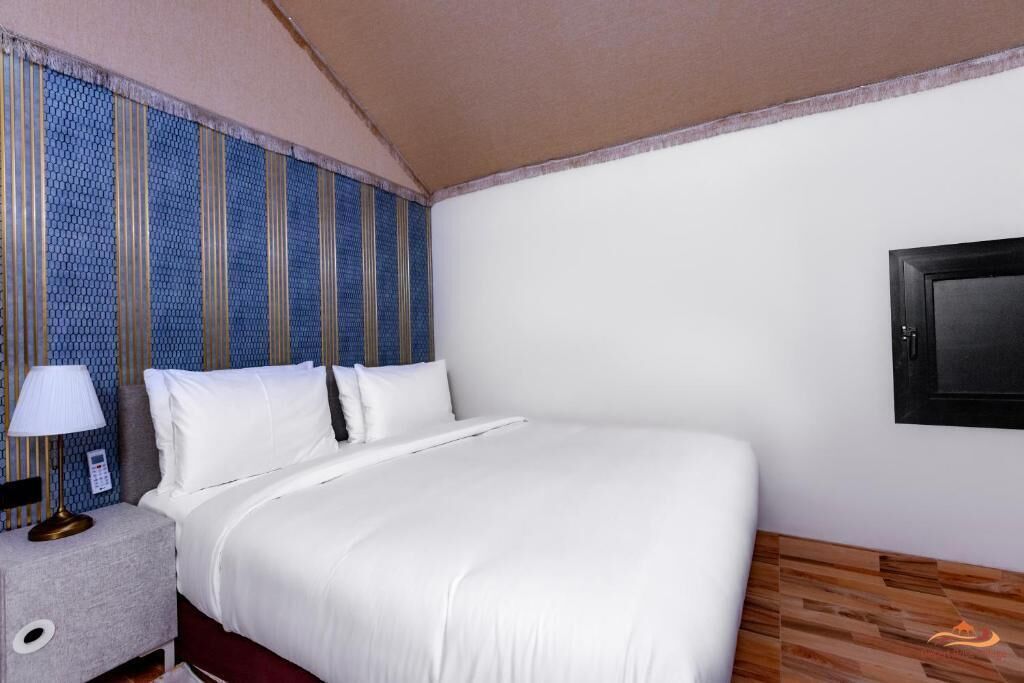 A hotel room with a bed, a nightstand with a lamp, a TV mounted on the wall, and a patterned wall.