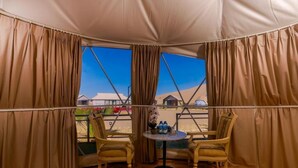 A tented room with two wooden chairs and a small table, a window with curtains, and a view of a desert landscape. - Desert Rose Village  (Ramlat Sweihan)