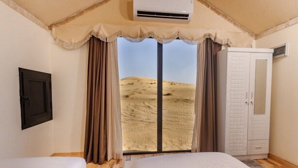 A bedroom with two beds, a wardrobe, an air conditioning unit, and a view of sand dunes through a large window. - Desert Rose Village  (Ramlat Sweihan)
