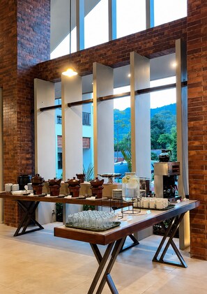Ein Buffet mit verschiedenen Speisen und einer Kaffeemaschine, vor einer Ziegelwand mit großen Fenstern.